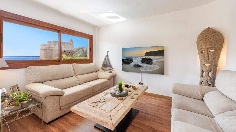 Sala de estar luminosa de villa mediterránea en Alcanada con vista al mar, suelo de madera y escultura de madera decorativa.