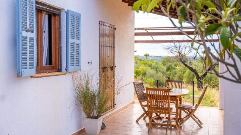 Covered terrace of the finca with wooden furniture, blue shutters and view into the green hinterland