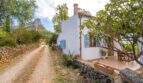 Exterior view of a white finca with blue shutters, gravel path, stone walls and rock massif in the background.