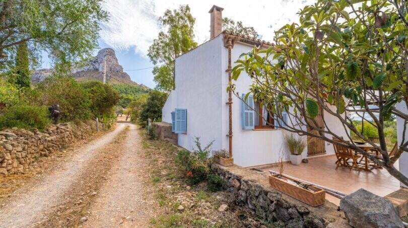 Exterior view of a white finca with blue shutters, gravel path, stone walls and rock massif in the background.