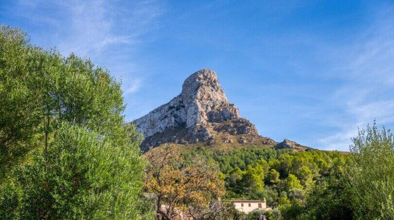 View of the distinctive rock peak with lush vegetation and traditional country house of the historic finca in Colònia de Sant Pere