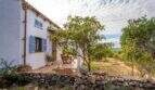 Exterior view of a historic finca with white facade, blue shutters and natural stone terrace surrounded by trees.