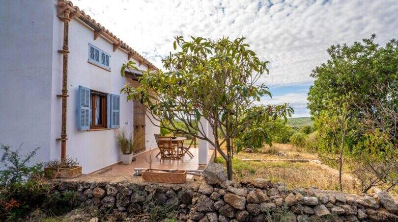 Exterior view of a historic finca with white facade, blue shutters and natural stone terrace surrounded by trees.