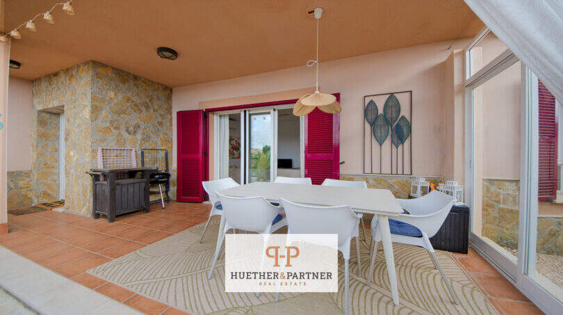 Überdachte Terrasse mit weißen Gartenmöbeln, Natursteinwand und pinken Fensterläden eines Hauses in Cala Murada