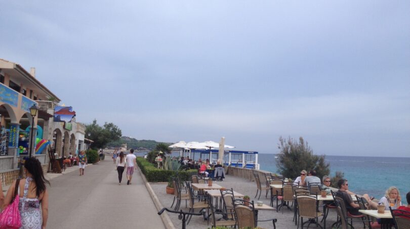 Strandpromenade in Cala Ratjada mit Cafés, Meerblick und türkisblauem Wasser direkt am Hafen.