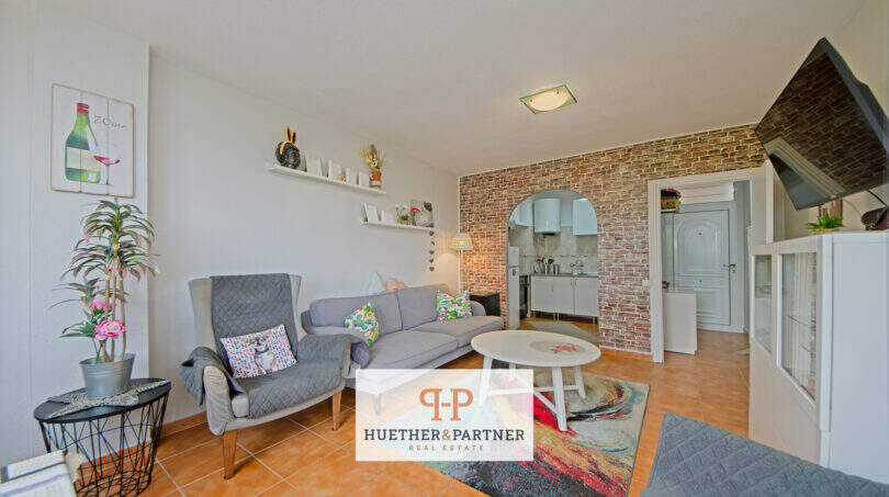 Helles Wohnzimmer der Mallorca-Wohnung mit Ziegelsteinbogen, grauer Couch und offener Küche.