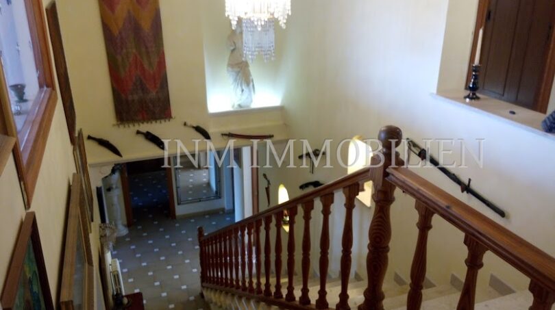 Elegante escalera con barandilla de madera, lámpara de araña y tapiz de pared en casa unifamiliar Santa Ponsa