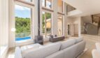 Bright living room of the villa in Cala Vinyes with high ceilings, fireplace, sofa and pool view through large windows.