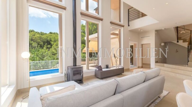 Bright living room of the villa in Cala Vinyes with high ceilings, fireplace, sofa and pool view through large windows.