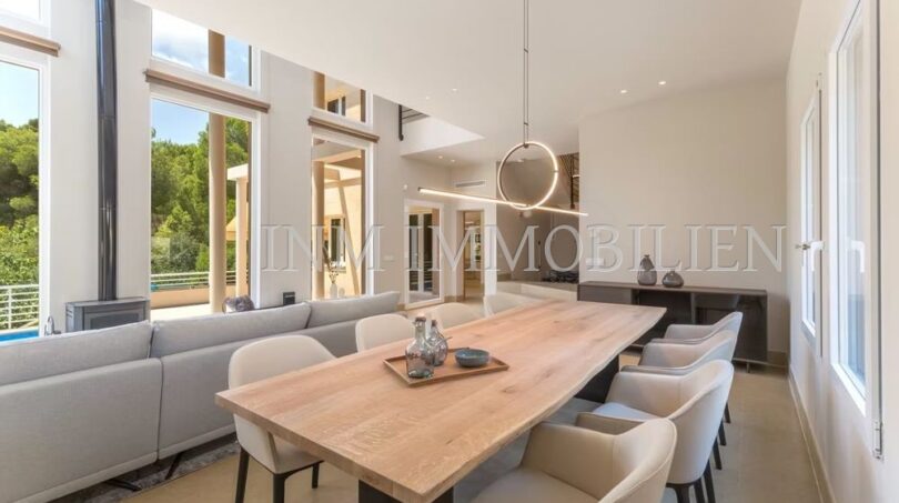Light-filled dining area with large wooden table, modern chairs and panoramic windows to the pool area of the villa in Cala Vinyes.