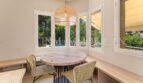 Bright dining room with round wooden table, rattan chairs and panoramic windows overlooking pool and garden of the villa.