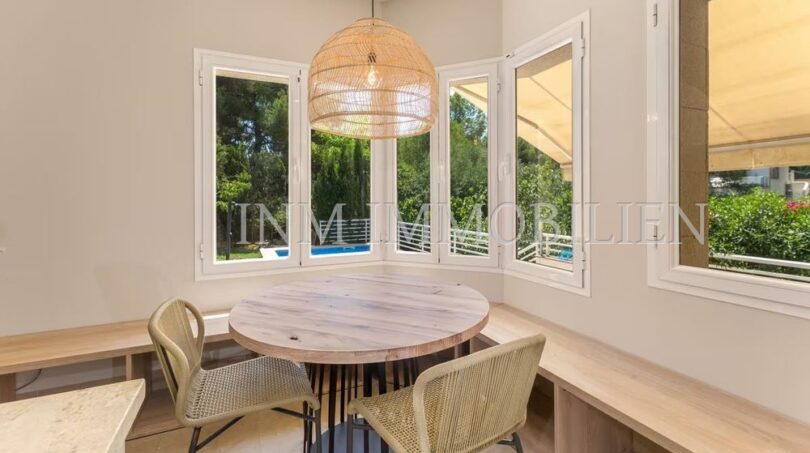 Bright dining room with round wooden table, rattan chairs and panoramic windows overlooking pool and garden of the villa.
