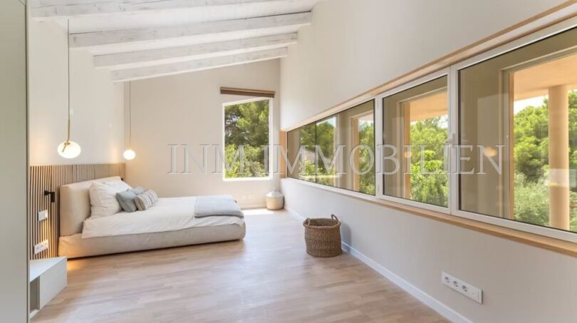 Bright bedroom of the villa in Cala Vinyes with wooden ceiling, parquet floor and panoramic window to the greenery.