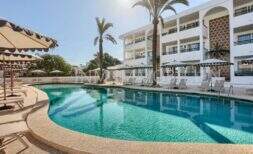 Pool area with lounge chairs and sun umbrellas in front of white hotel building with palm trees in Portocolom