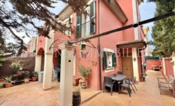 Vista exterior: Casa pareada en esquina roja bien cuidada con terraza cubierta, muebles de jardín y plantas en Pollença.