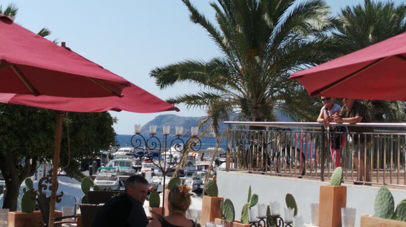 Hafenterrasse mit roten Sonnenschirmen, Palmen und Meerblick in Cala Ratjada, Mallorca
