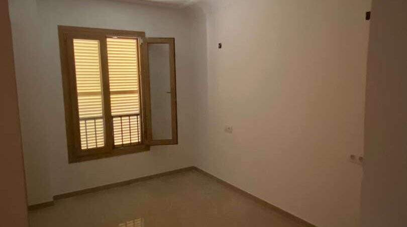 Leeres Zimmer der Hafenwohnung Porto Cristo mit Fliesenboden, weißen Wänden und Holzfenster mit Jalousie