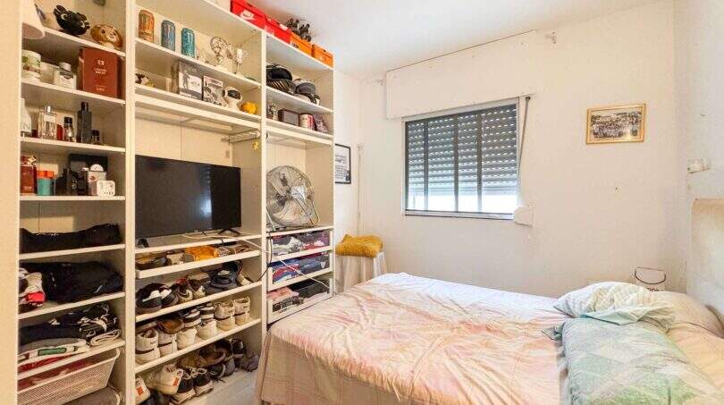 Kleines Schlafzimmer mit Einzelbett, weißen Regalen voller Schuhe und Elektronik sowie Fenster mit Jalousien