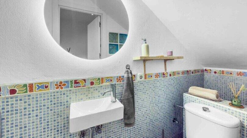 Modernes Badezimmer mit blauem Mosaikfliesen, buntem Fliesenbordüre, rundem LED-Spiegel und Wandwaschbecken im Stadthaus Felanitx