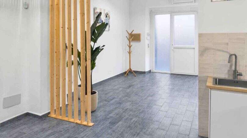 Modernes Wohnzimmer mit Holzraumteiler, Grünpflanze, grauem Fliesenboden und Miniküche in renoviertem Stadthaus Felanitx