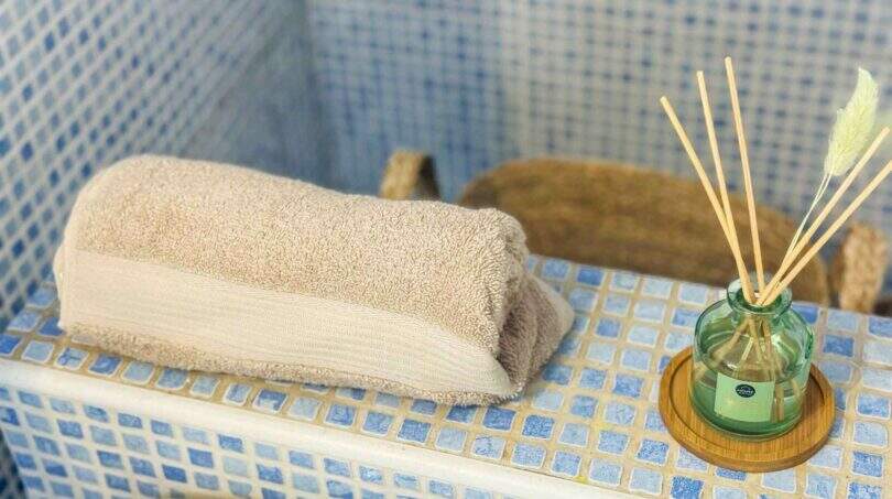 Badezimmer mit blauem Mosaikfliesenspiegel, gefalteten Handtüchern und Raumduft-Diffuser in grünem Glasfläschchen.