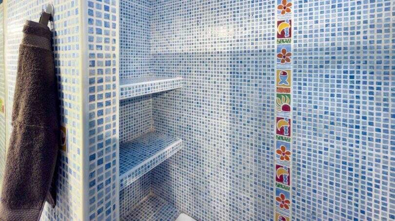 Dusche mit blauen Mosaikfliesen, buntem Fliesenband, zwei Einbauregalen und grauem Handtuch im Stadthaus Felanitx