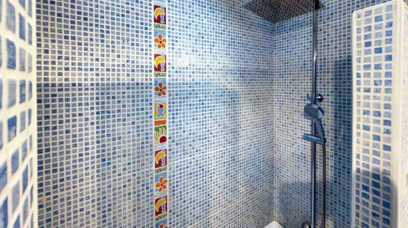 Badezimmer mit blauem Mosaikfliesenmuster, buntem Dekorstreifen und Duscharmatur über Badewanne