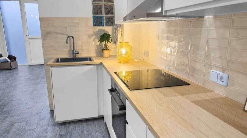 Moderne Küche mit Holzarbeitsplatte, Glaskeramikkochfeld, Einbauofen und beigen Wandfliesen im renovierten Stadthaus Felanitx