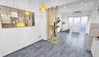 Helles Wohnzimmer mit grauem Vinylboden, Holzraumteiler, Hängeleuchte und Spiegelfenster zur Küche