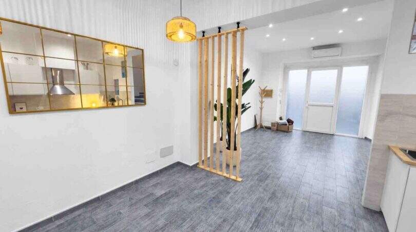 Helles Wohnzimmer mit grauem Vinylboden, Holzraumteiler, Hängeleuchte und Spiegelfenster zur Küche