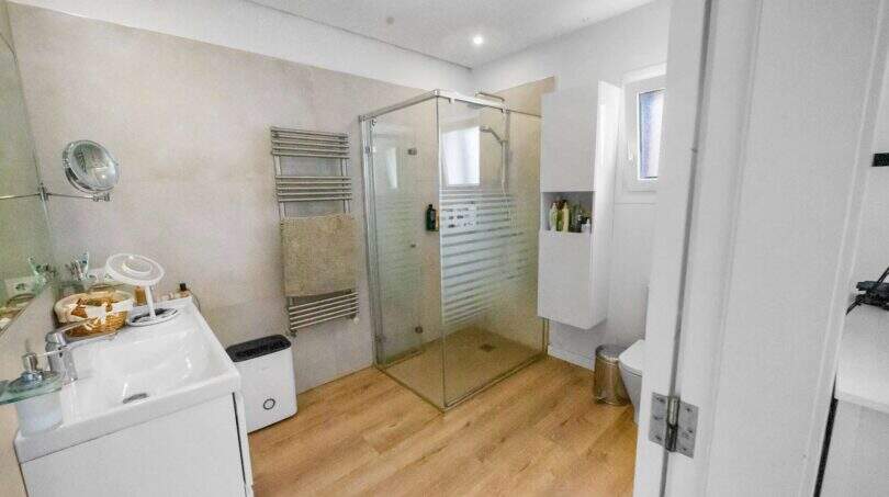 Modern bathroom with glass shower, wooden floor, towel radiator and white built-in furniture in house Felanitx