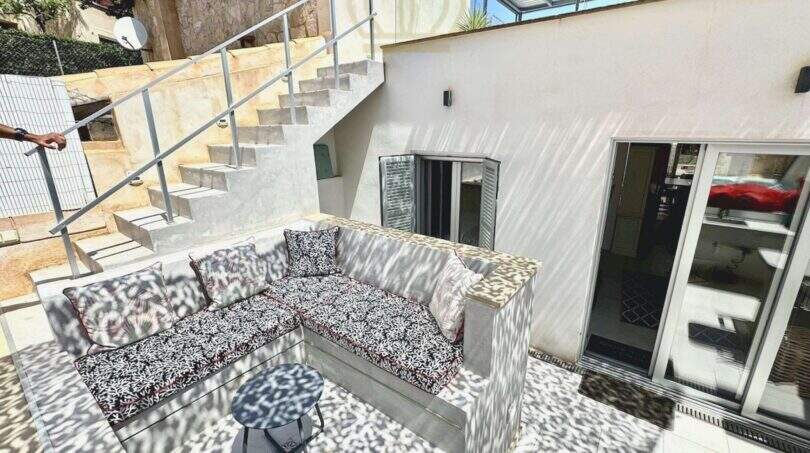 Sonnige Terrasse mit gemustertem Sofa, Betontreppe und Glastüren in saniertem Haus in Cala Llombards