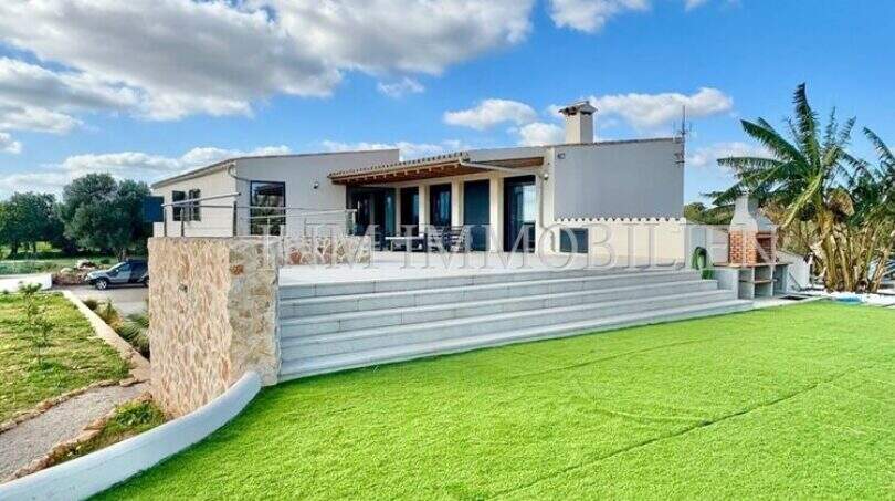 Exterior view Campos finca: modern country house with extensive lawn, terrace, stone wall and palm tree under blue sky.