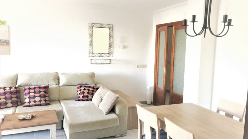 Helles Wohnzimmer der Mietwohnung in Cala Ratjada mit Ecksofa, Esstisch und elegantem Kronleuchter.