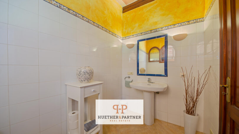 Helles Badezimmer der Finca Felanitx mit gelbem Deckenakzent, blauem Spiegel und weißen Wandfliesen.