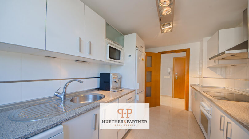 Moderne weiße Küche der Mallorca-Wohnung mit Granitarbeitsplatte, Einbaugeräten und hellem Fliesenboden.