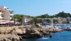 Hafenpromenade Cala Ratjada mit Booten, Felsen, Palmen und Gebäuden direkt am türkisblauen Meer