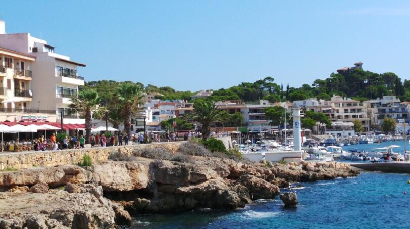 Hafenpromenade Cala Ratjada mit Booten, Felsen, Palmen und Gebäuden direkt am türkisblauen Meer