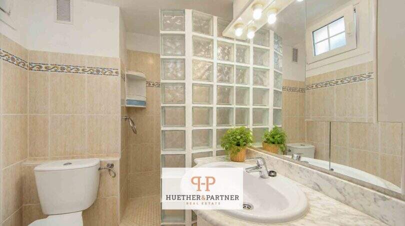 Helles Badezimmer der Mallorca-Wohnung mit Glasbausteindusche, Marmor-Waschtisch und beigen Fliesen.