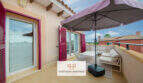Sonnige Dachterrasse mit Sonnenschirm, weißen Loungermöbeln und pinken Fensterläden in Cala Murada