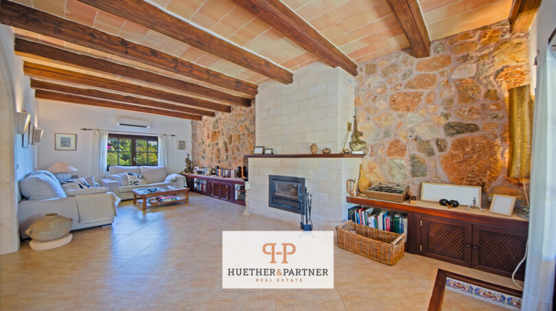 Wohnzimmer der Finca Felanitx mit Natursteinwand, Kamin und rustikalen Holzbalken an der Decke.