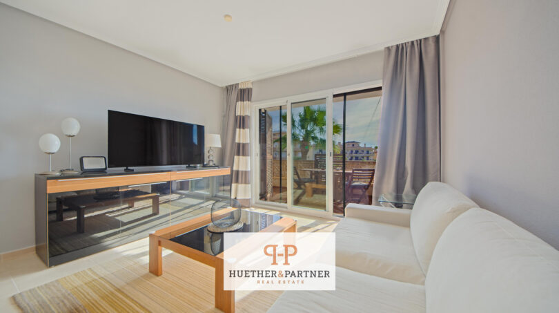 Helles Wohnzimmer der Mallorca-Wohnung mit Sofa, TV und Glasschiebetür zum sonnigen Balkon mit Palmenblick.