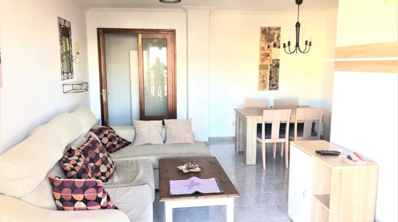 Helles Wohnzimmer der Mietwohnung in Cala Ratjada mit Ecksofa, Couchtisch und Essbereich mit Holztisch.