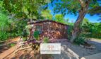 Casa de jardín de madera de la finca Felanitx, rodeada de árboles exuberantes y plantas verdes, con camino de grava y encanto rústico.