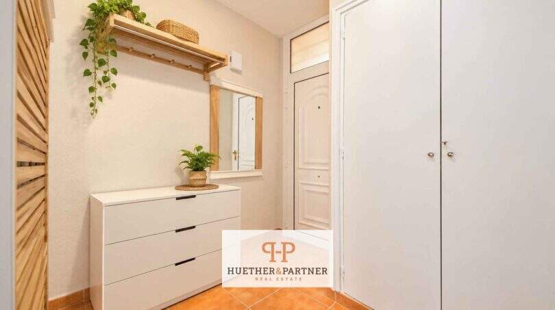 Heller Eingangsbereich der Mallorca-Wohnung mit weißem Kleiderschrank, Kommode und Holzregal mit Pflanzen.