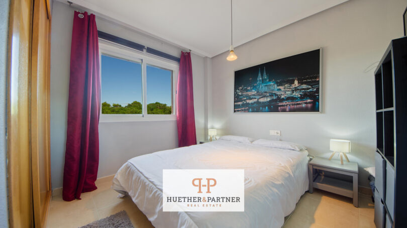 Helles Schlafzimmer der Mallorca-Wohnung mit Doppelbett, roten Vorhängen und Grünblick aus dem Fenster.