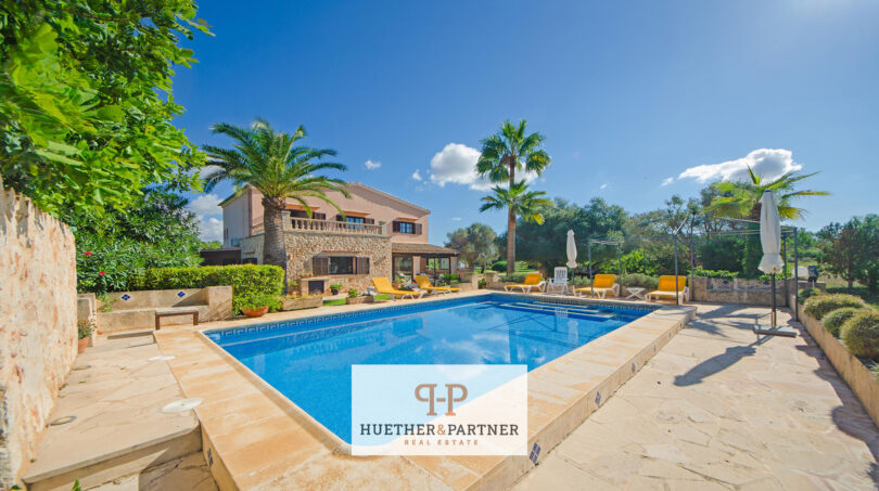 Finca Felanitx: Piscina mediterránea con terraza de piedra natural, palmeras y casa de campo rústica bajo cielo azul