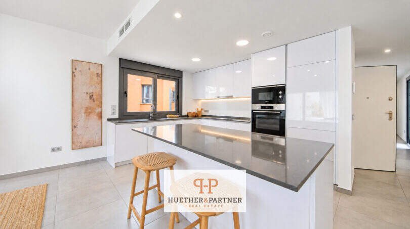 Moderne weiße Einbauküche mit Kochinsel, dunkler Granitarbeitsplatte und Barhockern im Penthouse Colonia de Sant Jordi