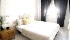 Luminoso dormitorio con cama doble, cojín decorativo amarillo, mesilla de noche de madera y cortinas oscuras cerca del mar, Alcudia