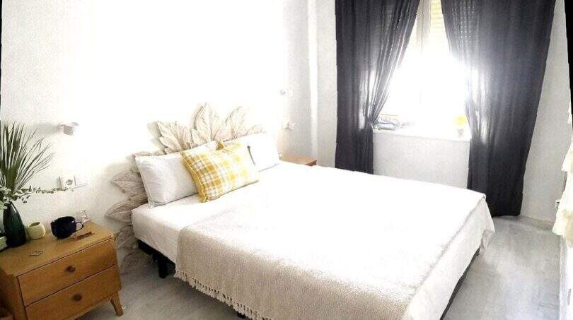 Luminoso dormitorio con cama doble, cojín decorativo amarillo, mesilla de noche de madera y cortinas oscuras cerca del mar, Alcudia